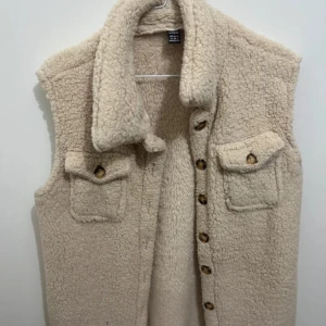 Beige pälsväst med knappar - Mysig beige pälsväst med knappar framtill och två fickor. Perfekt för kyliga dagar.