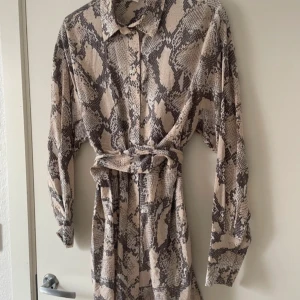 Beige och brun mönstrad klänning från H&M - Passar storlekar XS,S och M. I perfekt skick. En beige och brun klänning med ormmönster från H&M. Klänningen har långa ärmar och ett knytskärp i midjan för en snygg passform. Perfekt för en trendig look!
