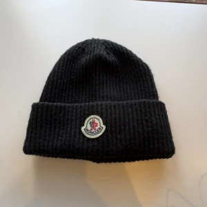 Svart mössa från Moncler - Snygg svart ribbad mössa från Moncler med deras ikoniska logotyp framtill. Perfekt för att hålla värmen under kyliga dagar. En stilren accessoar som passar till det mesta.