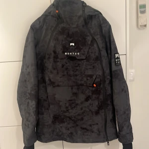 Svart jacka från Montec - Säljer en svart jacka från Montec med en cool mönstrad design. Jackan har en stor ficka framtill med Montec-logga och justerbara remmar. Perfekt för kyliga dagar med sin långa ärmar och huva.