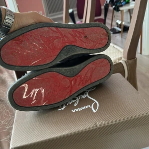 Lou Spikes Orlato Flat från Christian Louboutin - Säljer ett par grå sneakers från Christian Louboutin med spikdetaljer på tån och sidorna. Skorna har en ikonisk röd sula och snörning. Perfekta för den som vill sticka ut med stil.