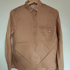 Beige långärmad skjorta med knappar - Säljer en stilren beige skjorta med långa ärmar och klassisk krage. Skjortan har en bröstficka med knapp och knäppning framtill. Perfekt för en avslappnad men ändå uppklädd look.