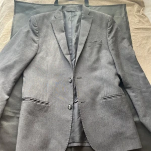 Grå kavaj från J.Lindeberg - Snygg grå kavaj från J.Lindeberg i slim fit. Perfekt för en stilren look. Kavajen har två knappar framtill och klassiska fickor. Tillverkad i högkvalitativt material för en elegant känsla.