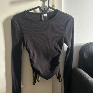 Svart ribbad topp från Divided - Säljer en svart ribbad topp från Divided med långa ärmar och snörning i sidorna. Perfekt för en stilren look. Toppen är i storlek XS och har en tight passform.