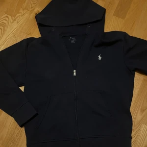 Svart hoodie från Polo Ralph Lauren - Säljer en stilren svart hoodie från Polo Ralph Lauren med dragkedja och klassisk logga på bröstet. Perfekt för en avslappnad look. Hoodien har långa ärmar och en praktisk huva.