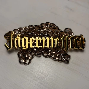 Jägermeister halsband - Snyggt halsband med ett stort guldfärgat Jägermeister emblem. Kedjan är robust och ger en cool look. Perfekt för att sticka ut på festen eller som en unik accessoar i vardagen.