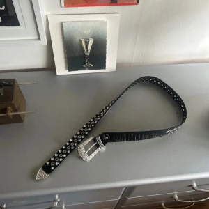 Svart bälte med nitar och silverdetaljer - Snyggt svart bälte med silverfärgade nitar och ett glittrande spänne. Bältet har en krokodilpräglad textur och spetsiga ändar med extra detaljer. Perfekt för att ge en edgy touch till din outfit.