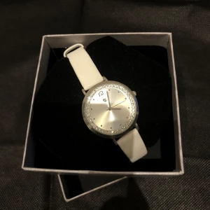 Vit klocka med glittrig urtavla - Elegant klocka med vit skinnarmband och en glittrig urtavla. Klockan har en silverfärgad boett och stilrena detaljer. Perfekt för att ge en touch av glamour till din outfit.