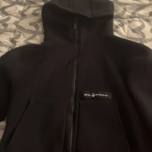 Svart tröja från Sail Racing M - Svart tröja från Sail Racing i storlek medium. Tröjan har huva, hel dragkedja framtill och två stora fickor. Snygg logga på bröstet och sportig look som passar perfekt till streetwear. Materialet känns slitstarkt och jackan är riktigt clean.