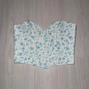 Blommig korsett från Zara i blått och vitt - Supersöt korsett från Zara med blått blommönster på vit botten. Toppen har en hjärtformad urringning och stängs med hyskor bak. Materialet är mjukt och lätt, perfekt för att skapa en romantisk look. Köpte den flera år sen men har aldrig använt. 