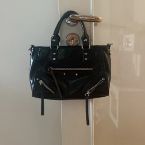 Svart handväska med dragkedjor - Snygg svart handväska i glansigt skinn med coola silverfärgade dragkedjor och nitar framtill. Väskan har två handtag och flera fack med dragkedja, perfekt för att hålla ordning på dina grejer. Stilren och edgy design som lyfter vilken outfit som helst.