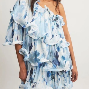 Blåvit blommig omlottklänning med volang - Superfin kortklänning i blått och vitt med blommigt mönster och stora volangdetaljer. Klänningen har en axel och ärmlös design på ena sidan, vilket ger en unik och trendig look. Perfekt för dig som vill sticka ut med en romantisk vibe.