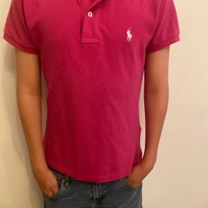 Rosa pikétröja från Polo Ralph Lauren - Snygg rosa pikétröja från Polo Ralph Lauren med klassisk krage och två knappar framtill. Tröjan har korta ärmar och den ikoniska vita loggan broderad på bröstet. Tillverkad i mjuk bomull som känns skön mot huden. Perfekt för en stilren och avslappnad look. Den är figursydd och passar både tjejer och killar. 