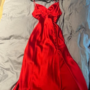 Röd långklänning i satin med knappar - Snygg röd långklänning i glansigt satinmaterial med smala axelband och v-ringad urringning. Klänningen har dekorativa knappar längs ena sidan och en slits som ger en extra touch. Perfekt för dig som vill sticka ut med en elegant och trendig look.