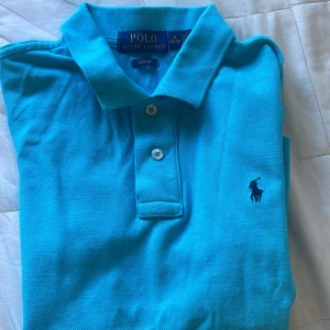 Blå pikétröja från Ralph Lauren - Snygg blå pikétröja från Ralph Lauren i slim fit-modell. Klassisk krage, två knappar och den ikoniska broderade loggan på bröstet. Perfekt för dig som gillar stilrena och sportiga plagg. Tillverkad i bomull för en skön känsla.