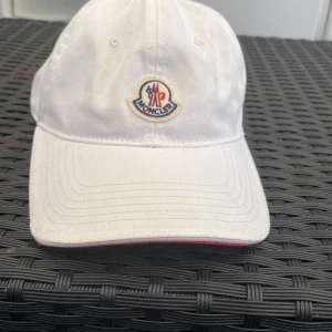 Vit keps från Moncler med logga - Snygg vit keps från Moncler med klassisk böjd skärm och broderad logga framtill. Justerbar rem baktill i rött, vitt och blått med metallspänne. Kepsen är i bomull och har röda detaljer på insidan för extra stilpoäng.