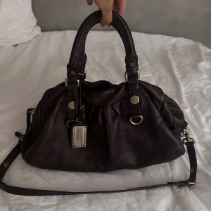 Marc Jacobs väska - Säljer en mörkbrun handväska från Marc Jacobs i modellen Classiq Q baby groove💕💕obs den har ett hål i innerfickan (men inget som syns). Har varit en favorit!!🥹