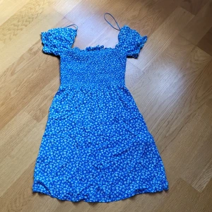Blå blommig kortklänning New yoker - Supersöt blå kortklänning från FB Sister med vita små blommor. Klänningen har puffärmar, smockad överdel och tunna axelband. Perfekt för sommarens varma dagar och ger en riktigt fräsch och ungdomlig vibe.