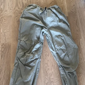 Gråa parachute byxor från Divided M - Säljer ett par gråa parachute byxor  från Divided i storlek M. Byxorna har resår och snörning i midjan samt stora fickor på sidorna. De är tillverkade i bomull och har en loose fit som ger en avslappnad vibe.