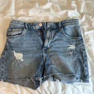 Blå jeansshorts med slitningar H&M - Snygga blå jeansshorts från H&M med slitna detaljer och fransig kant nertill. Klassisk femficksmodell med dragkedja och knapp. Perfekta för sommaren och ger en avslappnad vibe till din outfit.