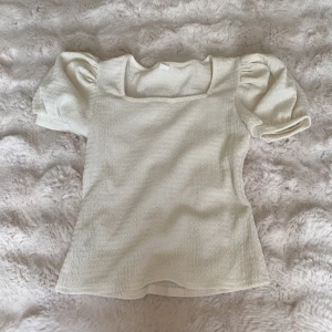 Vit puffärmad blus med struktur - Supersöt vit blus med puffiga korta ärmar och fyrkantig ringning. Tyget har en vågig struktur som ger en unik känsla. Perfekt att styla med jeans eller kjol för en trendig look.