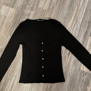 Svart ribbad kofta från Zara - Svart ribbad kofta från Zara med långa ärmar, v-ringning och dekorativa spetsdetaljer framtill. Snygga vita knappar längs hela framsidan ger en clean kontrast. Perfekt att styla med jeans eller kjol för en trendig look. Knapparna går ej att knäppa upp! Storlek M men är lite liten i storlek och passar mig som har S vanligtvis🩷. Andvänd en gång