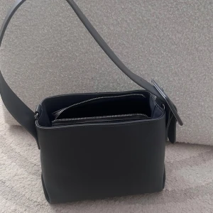 Svart axelväska med bredt spänne - Stilren dammblå/grå i polyuretan med bred axelrem och stort fyrkantigt spänne. Väskan har en rektangulär form, dragkedja upptill och medföljande lång rem för crossbody. Perfekt för dig som gillar minimalistisk design och vill ha plats för det viktigaste.