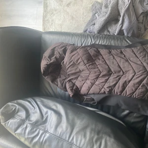 Svart dunväst med huva och dragkedja - Säljer en svart dunväst med snyggt chevronmönster och hel dragkedja framtill. Västen har en praktisk huva med dragsko och är perfekt för lager-på-lager. Materialet är mjukt och quiltat för extra värme. Passar dig som vill ha en stilren och funktionell look. Ny pris 500