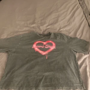 Grå t-shirt Love Sick från Primark - Cool grå t-shirt från Primark i bomull med tryck av ett spraymålat rosa hjärta och texten 'Love Sick' på bröstet. Klassisk passform och korta ärmar, perfekt för en edgy streetstyle-look.