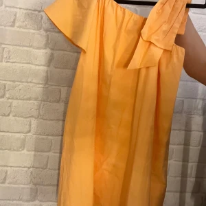 Orange offshoulder klänning med rosett - Superfin orange kortklänning i bomull med offshoulder-design och en stor rosett på ena axeln. Klänningen har en lös passform och är perfekt för varma dagar. Enkel och stilren utan mönster, med en fräsch och somrig vibe. Aldrig använd💗