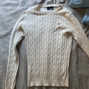 Vit kabelstickad tröja från Gant - Kabelstickad vit tröja från Gant med rund halsringning och diskret logga på bröstet. Tröjan har lång ärm och klassisk passform, perfekt för lager-på-lager. Mjuk och skön känsla tack vare bomullsmaterialet.