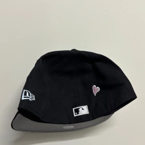 New era 59fifty keps  - Svart LA Dodgers keps från New Era 59FIFTY med broderad LA-logga framtill, palmer och hjärta på sidan samt Dodger Stadium 50th Anniversary-märke. Grå underskärm och klassisk flat peak. Perfekt för dig som vill sticka ut med detaljer.