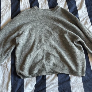 Grå stickad tröja med rund hals - Mysig grå stickad tröja med rund halsringning och långa ärmar. Tröjan har en relaxed passform och ribbade muddar vid ärmslut och nederkant. Perfekt för lager-på-lager under kyliga dagar.