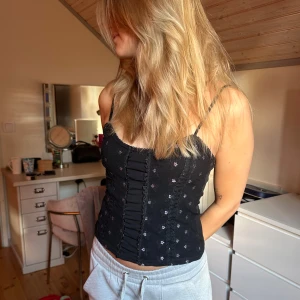 Svart blommig korsett-topp med spets - Supersöt svart korsett-topp med tunna axelband och små lila blommor. Framsidan har dekorativa spetsdetaljer och rynkade sömmar som ger en feminin vibe💜