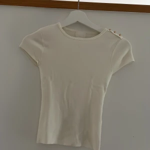 Vit ribbad topp med guldknappar H&M XS - Superfin vit ribbad topp från H&M i storlek XS. Toppen har korta ärmar och rund halsringning, samt tre snygga guldiga knappar på ena axeln som cool detalj. Perfekt att styla med jeans eller kjol för en clean look. Orginal pris 200kr, priser kan diskuteras.