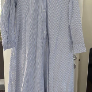 Blåvit randig skjortklänning från Zara - Snygg lång skjortklänning från Zara i blått och vitt med tunna svarta ränder. Klänningen har knappar hela vägen framtill och på sidorna, klassisk skjortkrage och långa ärmar. Perfekt för en chill och stilren look.