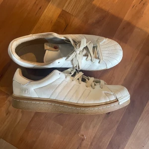 Vita Adidas Superstar sneakers i skinn - Adidas Superstar sneakers i vitt skinn med klassisk snäcktå och beige sula. Snygga detaljer med broderad '1997' på sidan och ikoniska tre ränder. Skorna har snörning och en platt sula, perfekta för en clean streetstyle-look.