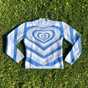 Blå heartwave tröja - Superfin stickad tröja från Cider i blått och vitt med stora hjärtmönster på både fram- och baksidan. Tröjan har lång ärm, rund hals och är gjord i en mjuk mix av bomull och akryl. Perfekt för dig som gillar färg och mönster! Tröjan är använd, vilket syns på små noppror och en tråd som dragits lös på ärmen