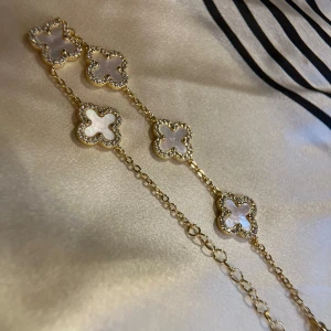 Guldigt halsband med vita fyrklöver - Superfint halsband i guldton med flera fyrklöverformade hängen i vit pärlemor och glittrande stenar runtom. Kedjan är tunn och elegant med justerbar längd. Perfekt för dig som vill ha något som sticker ut och känns lyxigt. 21cm in total 