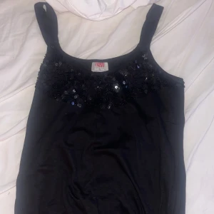 Svart paljettlinne från Flash - Snygg svart linnetopp från Flash med breda axelband och glittriga paljetter upptill. Perfekt för dig som vill sticka ut lite extra. Mjuk och bekväm, med en enkel men ändå festlig look. Storlek m men passar xs eller s
