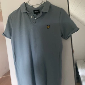 Ljusblå pikétröja från Lyle & Scott - Snygg ljusblå pikétröja från Lyle & Scott med klassisk krage och korta ärmar. Tröjan har den ikoniska gula loggan broderad på bröstet och är tillverkad i mjuk bomull. Perfekt för en clean och stilren look.