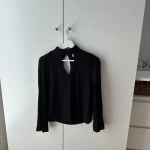 Svart blus med cut out och knappar - Svart blus i mjukt tyg med lång ärm och hög krage. Snygg cut out-detalj framtill och tre små knappar i nacken. Perfekt om du gillar stilrena plagg med en twist. Passar till både jeans och kjol.