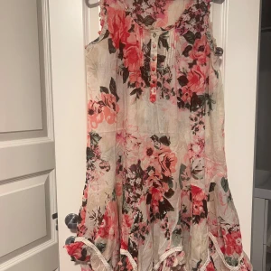 Blommig kortklänning från RoseBud - Supersöt kortklänning från RoseBud i 100% bomull med blommigt mönster i rosa, rött och grönt på ljus botten. Ärmlös modell med knappar framtill, volangdetaljer och romantisk känsla. Perfekt för sommarens alla tillfällen!