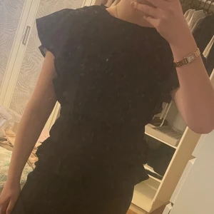 Svart volangklänning med kort ärm - Snygg svart klänning med volangdetaljer längs axlar och sidor. Modellen har korta ärmar och markerad midja, vilket ger en feminin siluett. Perfekt för dig som gillar stilrena och eleganta plagg.