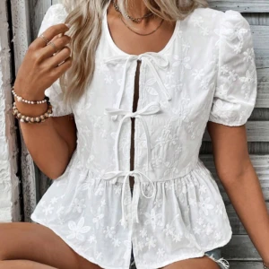 Vit broderad blus med knyt - Superfin vit blus med broderade blommor och tre knyt framtill. Puffiga korta ärmar och lätt utsvängd nertill ger en söt och romantisk vibe. Perfekt till jeansshorts eller kjol för en fräsch look.