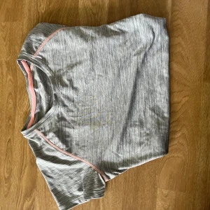 Grå sportig t-shirt med rosa detaljer - Snygg gråmelerad t-shirt med korta ärmar och rosa sömmar längs axlarna. Perfekt för träning eller chill dagar. Tillverkad i ett lätt och mjukt syntetmaterial som andas bra. Enkel och stilren design som passar till det mesta.