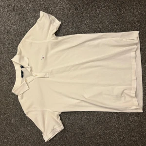Vit pikétröja från Gant - Vit pikétröja från Gant med klassisk krage och två knappar framtill. Liten broderad logga på bröstet. Skön och stilren t-shirt i bomull, perfekt till jeans eller shorts. Storleken är 176, passar både XS och S