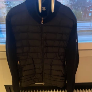 Svart Moncler cardigan med dragkedja - Snygg svart cardigan från Moncler med quiltad front och släta ärmar. Jackan har dragkedja framtill, två sidofickor med dragkedja och Moncler-logga på ärmen. Perfekt för dig som vill ha en stilren och sportig look.