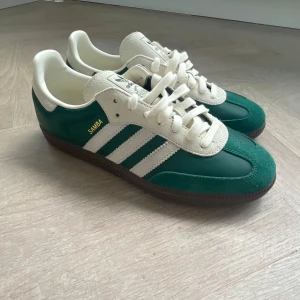 Adidas Samba grön/vit sneakers - Adidas Samba sneakers i grön och vit med klassiska tre ränder på sidan och guldfärgad 'SAMBA'-text. Ovandel i skinn och mocka, vit plös och snörning. Mörkbrun sula och retrovibe som passar perfekt till streetwear. Följer med mörkgröna snören. Tveka inte att höra av dig!!!
