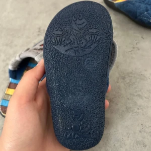 Blå tofflor med spindelbroderi Gezer - Mysiga blå tofflor från Gezer med quiltad ovandel i mjuk plysch och broderad röd spindel på varje sko. Insidan har färgglada ränder i gult, blått och brunt. Perfekta för att chilla hemma och hålla fötterna varma. Priset går att diskutera!